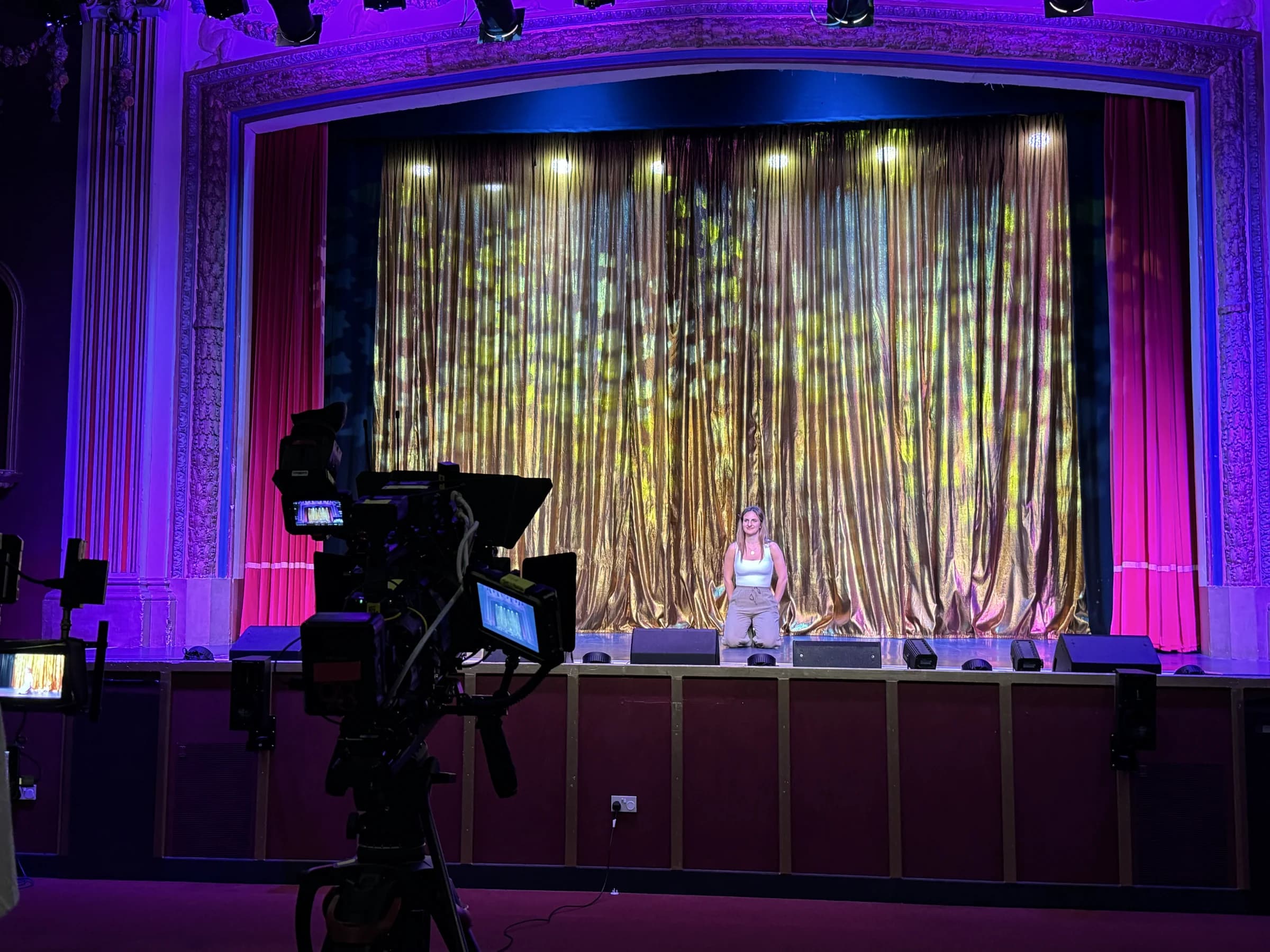 A broadcast camera framing a presenter at centre stage